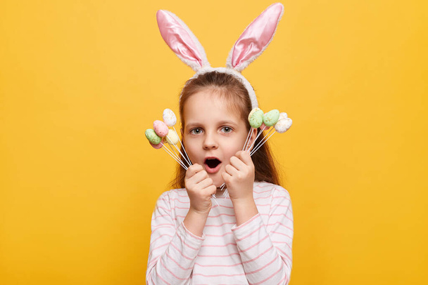 Portrait of excited scared surprised little girl with rabbit ears on her head posing isolated on yellow background, holding cake pops, keeps mouth open. - Foto, afbeelding