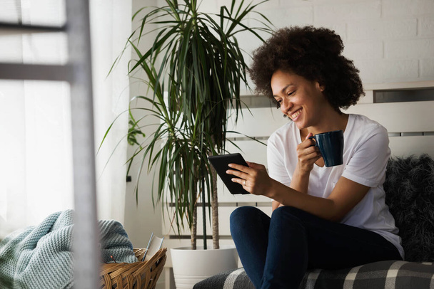 jovem afro-americana sentada em confortável sofá na sala de estar e usando tablet digital  - Foto, Imagem