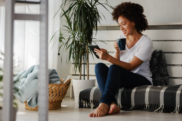молода афроамериканка сидить на зручному дивані у вітальні і користується цифровим планшетом.  - Фото, зображення