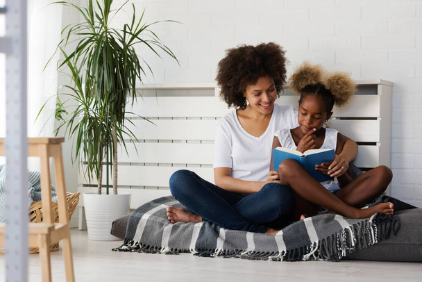 Όμορφη Αφρικανή Μητέρα και Κόρη διαβάζουν ένα βιβλίο μαζί στο σπίτι. Άνετη γωνία. - Φωτογραφία, εικόνα