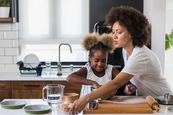 Мать учит ребенка готовить и помогать на кухне. Афроамериканская мать и дочь пекут печенье дома. - Фото, изображение
