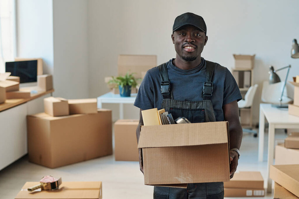 Portret Afroamerykanina uśmiechającego się do aparatu trzymającego pudełko z rzeczami, pomagającego przenieść się do nowego biura - Zdjęcie, obraz