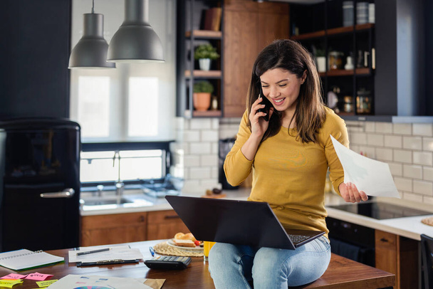 Młoda uśmiechnięta bizneswoman pracująca w domu, siedząca przed laptopem w kuchni domowej - Zdjęcie, obraz