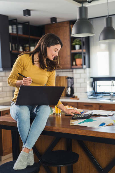 Junge lächelnde Geschäftsfrau, die von zu Hause aus arbeitet, sitzt zu Hause in der Küche mit Computer auf ihrem Laptop - Foto, Bild