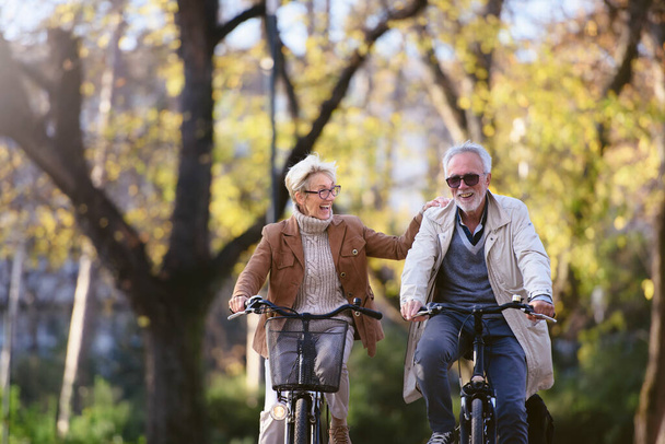 Cheerful active senior couple with bicycles in public park together having fun. Perfect activities for elderly people. Happy mature couple riding bicycles in park - Фото, изображение