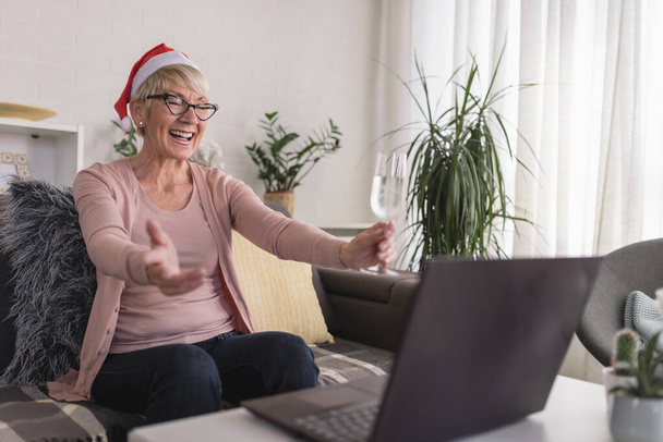 Eine alte Dame mit Nikolausmütze trinkt allein zu Hause Champagner und stößt auf einen Computer an. Videotelefonate mit der Familie im Urlaub über das Internet. - Foto, Bild