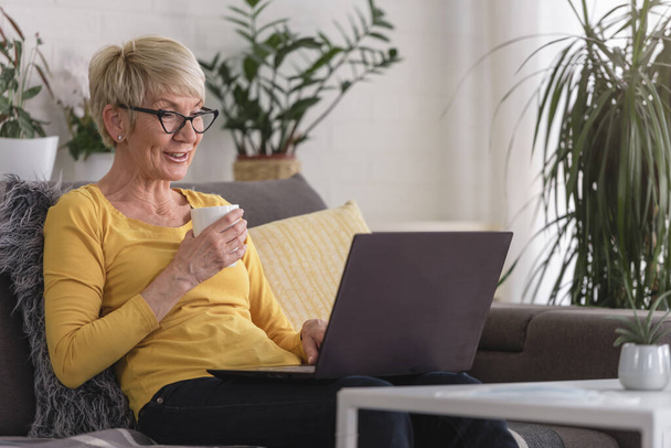 Όμορφη ηλικιωμένη γυναίκα στο σπίτι χρησιμοποιώντας φορητό υπολογιστή για να επικοινωνήσει με την οικογένειά της, σερφάροντας στο διαδίκτυο, ψωνίζοντας online, πίνοντας καφέ - Φωτογραφία, εικόνα