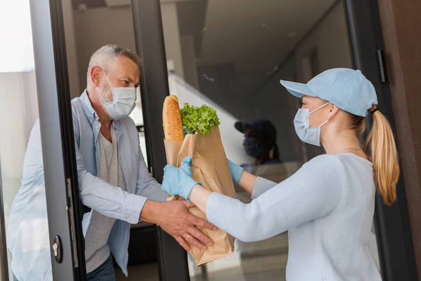 Девушка из службы доставки приносит продукты пожилому человеку во время пандемии COVID. люди в защитных масках. - Фото, изображение