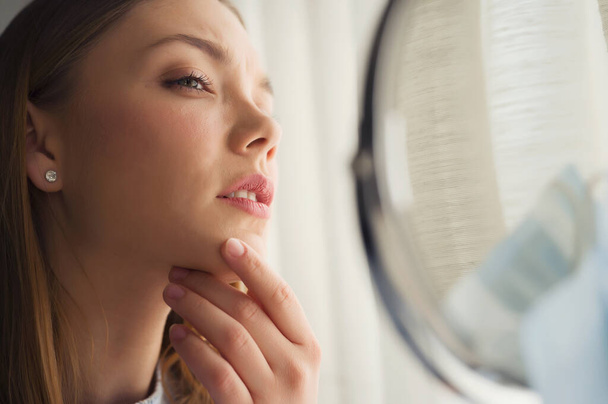 Jovem mulher bonita olhando-se no espelho em casa. Preocupado com acne. Problemas com acne. - Foto, Imagem