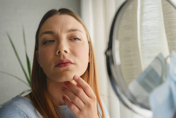 Young beautiful woman looking herself in the mirror at home. Worried about acne . Problems with acne. - Foto, afbeelding