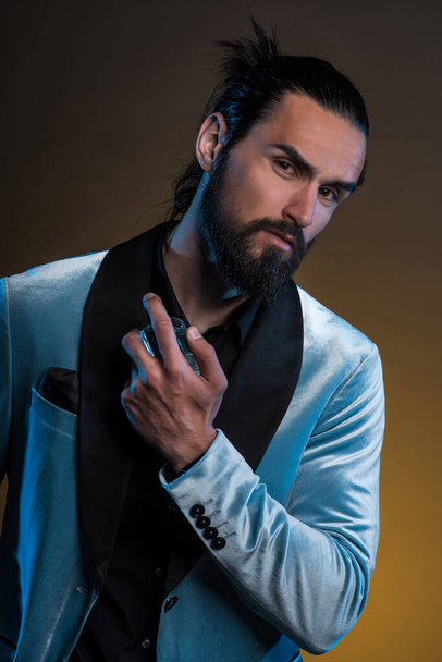 Beautiful masculine bearded young man holding a bottle of fragrance. Masculine perfume, bearded man in a suit. - Photo, Image
