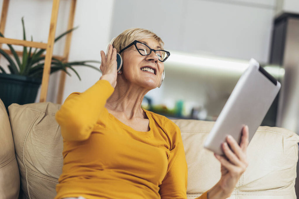 デジタルタブレットで笑顔ヘッドフォンで幸せな高齢者の女性と自宅で音楽を聴く - 写真・画像
