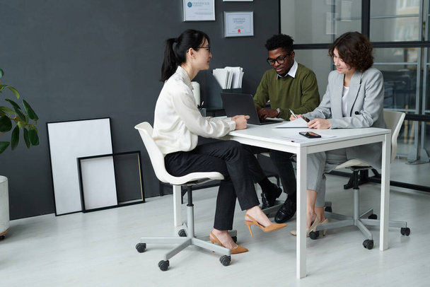 Молода жінка сидить за столом разом з менеджерами і розмовляє з ними під час співбесіди в сучасному офісі
 - Фото, зображення
