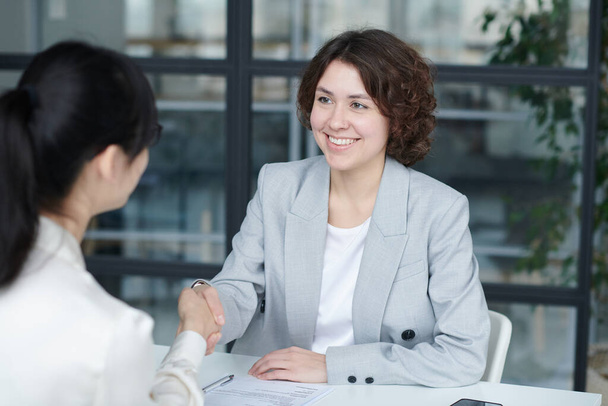 Jeune manager souriant serrant la main d'une employée pour la trouver du travail, ils sont assis à table lors d'une réunion d'affaires - Photo, image