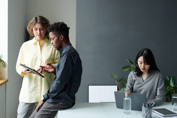 Dos colegas jóvenes discutiendo la presentación en línea juntos en la tableta digital con la mujer de negocios que trabaja en el ordenador portátil en segundo plano - Foto, imagen