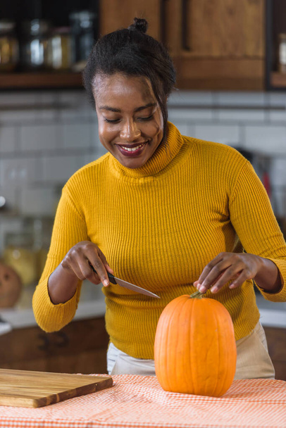 Giovane donna nera sorridente che si prepara per Halloween intagliando una zucca a casa in cucina - Foto, immagini