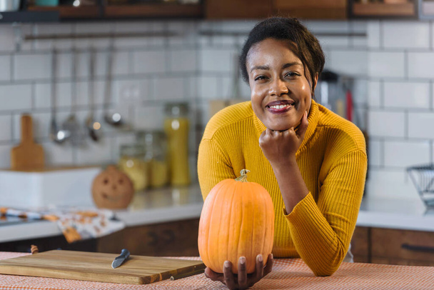 Young smiling black woman preparing for Halloween carving a pumpkin at home in the kitchen - Photo, Image