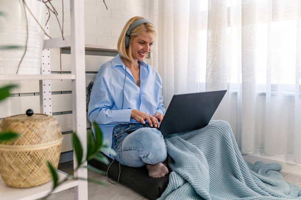 Молодая обычная женщина сидит у окна дома, используя ноутбук и наушники для общения через интернет. Интернет-музыка. Дистанционное обучение - Фото, изображение