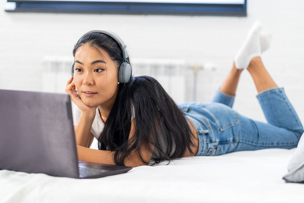 Asyalı bir öğrenci, evde internet üzerinden İngilizce öğrenen genç bir kadın. Kulaklık ve dizüstü bilgisayar kullanarak, online bir derse katılarak. - Fotoğraf, Görsel