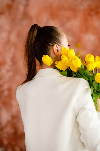 Una ragazza con dei tulipani gialli sulla spalla. Giacca bianca da donna. Acconciatura elegante. La festa della mamma sta arrivando presto - Foto, immagini