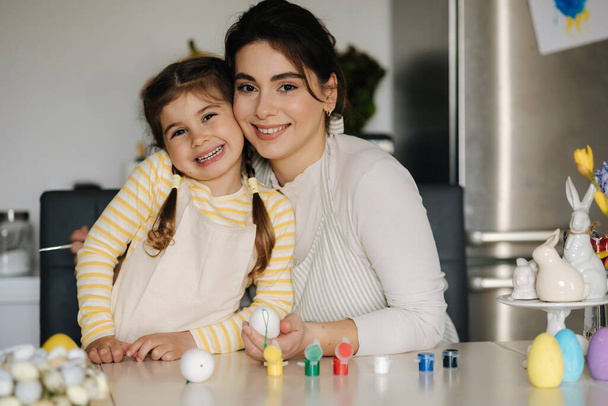 Portrait of adorable little girl with mom on kitchen. Beautiful woman help her daughter paint easter eggs. High quality photo - Foto, imagen