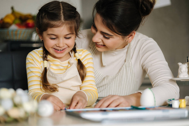 Šťastná, smějící se holčička maluje obrázek s mámou u stolu v kuchyni. Velikonoční prázdniny. Jaro. Kvalitní fotografie - Fotografie, Obrázek