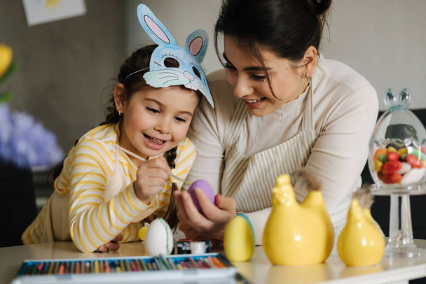 家族と一緒にイースターの時間。イースターバニーマスクの愛らしい女の子は、お母さんが卵を描くのを助ける。キッチンで春の装飾。高品質の写真 - 写真・画像