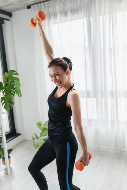 Lachende jonge vrouw aan het trainen met halters thuis. Dagelijkse lichaamsbeweging - Foto, afbeelding
