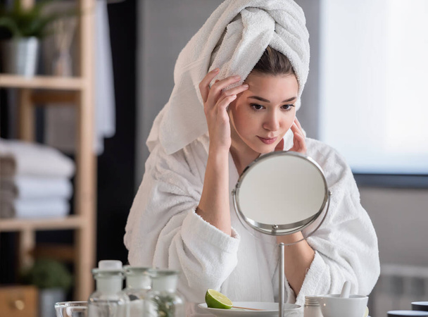 hermosa mujer joven en albornoz y toalla mirando al espejo, concepto de cuidado de la piel - Foto, Imagen