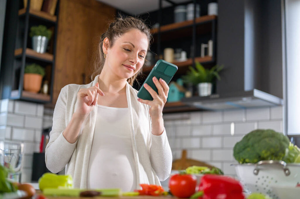 красивая беременная женщина с телефоном в руке, стоящая на кухне - Фото, изображение