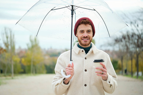 Junger Mann mit Kaffee und durchsichtigem Regenschirm im Winter auf der Straße. - Foto, Bild
