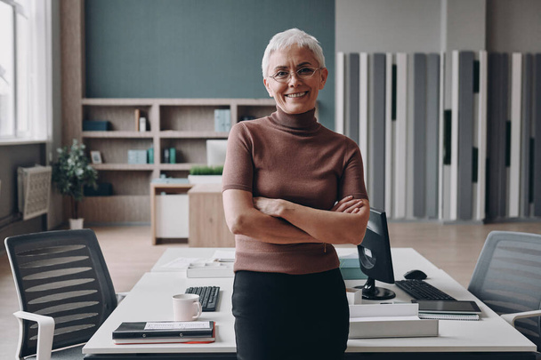 Senior businesswoman keeping arms crossed and smiling while leaning at the office desk - Foto, Imagem