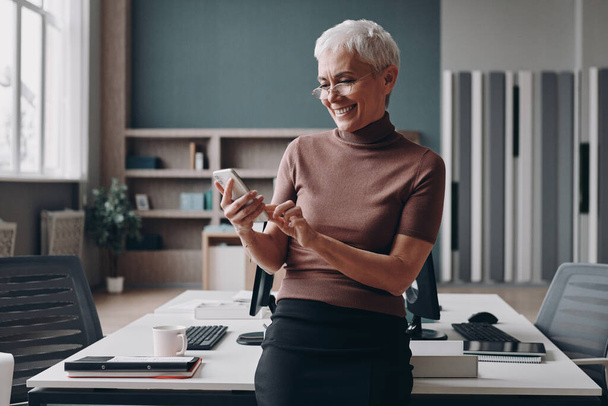 Senior businesswoman using smart phone and smiling while leaning at the office desk - Photo, Image