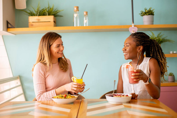 Two pretty young black and caucasian woman having good time, drinking fresh juices and having healthy breakfast in the cafe - Foto, afbeelding