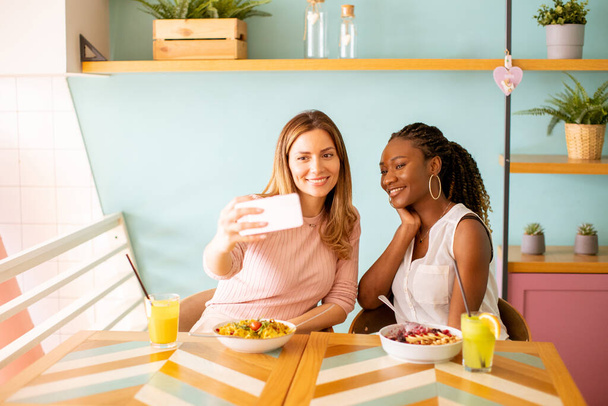 Zwei hübsche junge Frauen, kaukasisch und schwarz, machen Selfie mit Handy im Café - Foto, Bild