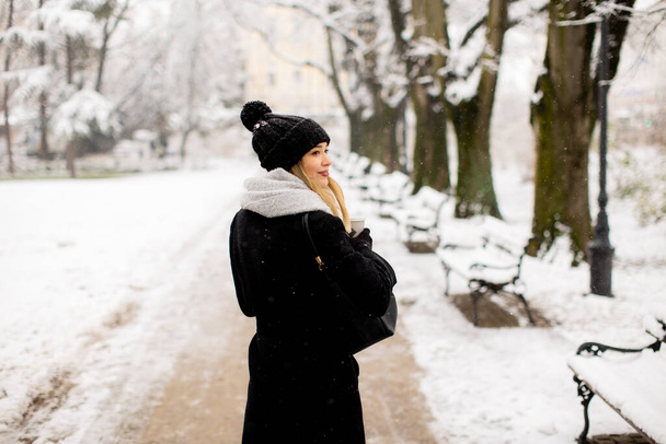Ładna młoda kobieta w ciepłych ubraniach korzystających w śniegu z kubka na wynos kawy - Zdjęcie, obraz