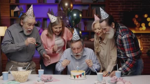 誕生日の帽子の面白い高齢者は、老人ホームで他の高齢者と一緒に祝う、ジョークの誕生日ケーキにキャンドルを吹いています。誕生日パーティー、古い友人がパーティーの角を吹いている - 映像、動画