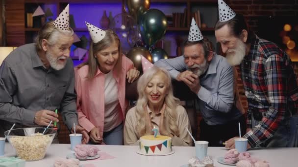 ブロンドは、退職家で他のシニアの人々と一緒に祝う、ジョークの誕生日ケーキにキャンドルを吹いて誕生日の帽子で高齢者の女性を微笑んだ。誕生日パーティー、古い友人がパーティーの角を吹いている - 映像、動画