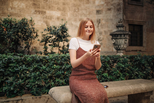 Chica señalando el dedo en el teléfono inteligente pantalla. Joven mujer elegante sonriente sosteniendo teléfono inteligente caminando en la calle al aire libre. Antiguo hermoso centro de Valencia España - Foto, imagen