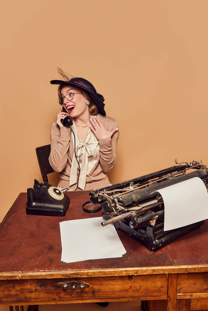 Good news. Charming woman sitting at table and talking on phone with pleasantly surprised face over beige background. Concept poems, novel, emotions, beauty, fashion, retro style, vintage - Foto, Imagen