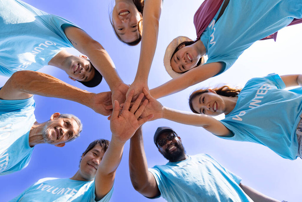 Equipo de jóvenes y el grupo de trabajadores voluntarios diversidad disfrutar de trabajo social caritativo al aire libre juntos en el ahorro de proyecto de medio ambiente vistiendo camiseta azul mientras se unen de la mano en el poder ensamblar la unidad - Foto, imagen