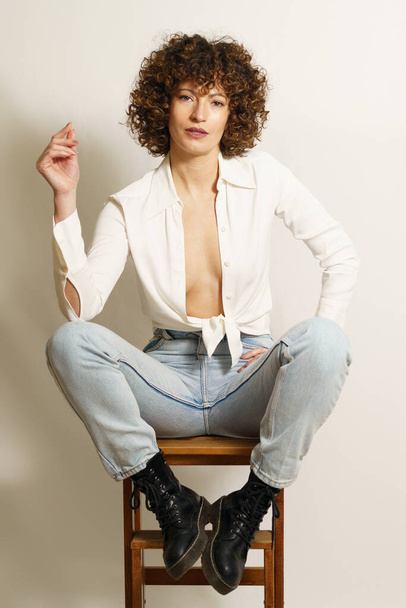 Full length of flirty female model with curly hair, in stylish wear resting on wooden stool playing with curl and looking at camera against white background - Photo, image
