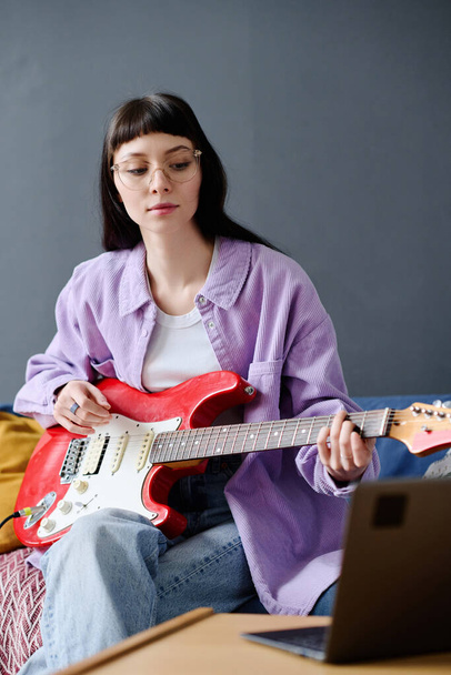 Молодая женщина, сосредоточенная на игре на гитаре, она смотрит на монитор ноутбука и учится играть - Фото, изображение
