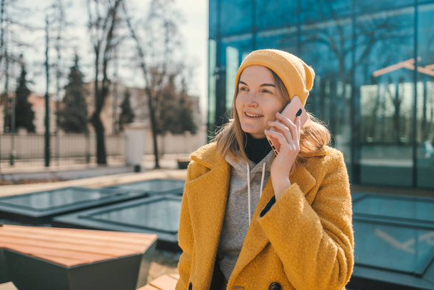 Horizontal shot of happy young woman makes telephone call wears casual sweatshirt and yellow coat had glad expression walks against blurred city building enjoys smartphone talking - Photo, Image