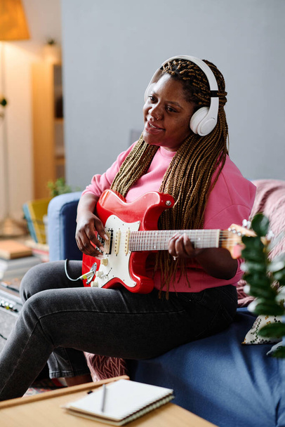 Africká americká dívka v bezdrátových sluchátkách hrát na kytaru a zpívat píseň, zatímco sedí na pohovce v místnosti - Fotografie, Obrázek