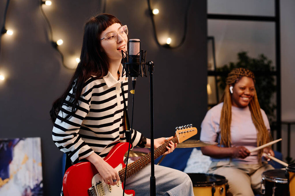 Young woman playing guitar and singing while rehearsing with her friend playing drums in background - 写真・画像