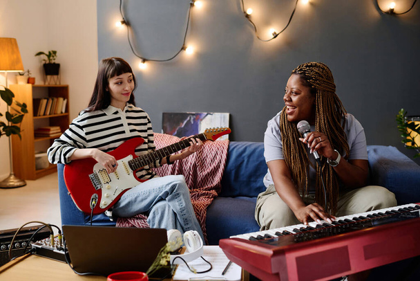 Δύο κορίτσια κάθονται στον καναπέ και παίζουν μουσική μαζί στο στούντιο χρησιμοποιώντας μικρόφωνο και μουσικά όργανα - Φωτογραφία, εικόνα