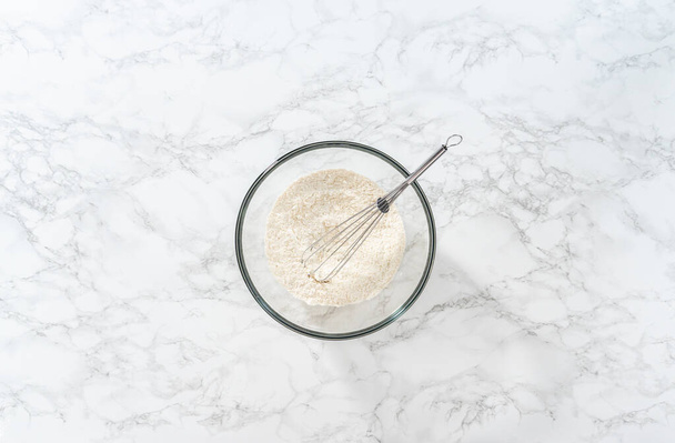 Flat lay. Mixing dry ingredients with a hand whisk in a large glass mixing bowl to bake mini vanilla cupcakes with ombre pink buttercream frosting. - Valokuva, kuva