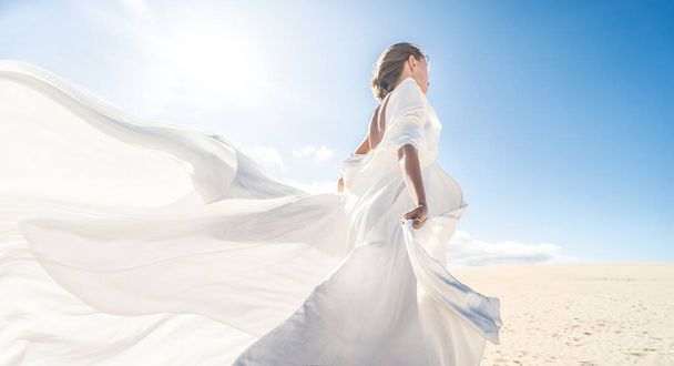 Photo of a woman in amazing white wedding dress posing on the desert, sunny light, blue sky, beautiful pastel tone, copy space. - Фото, изображение