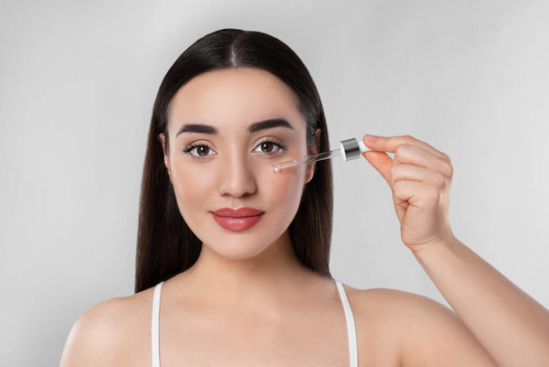 Young woman applying essential oil onto face on light grey background, closeup - Φωτογραφία, εικόνα